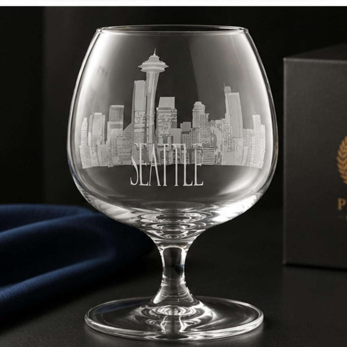 A clear glass cognac or brimman glass with an etched Seattle skyline and the word 'SEATTLE' on the bowl, shown on a dark surface with a dark box and blue fabric in the background.