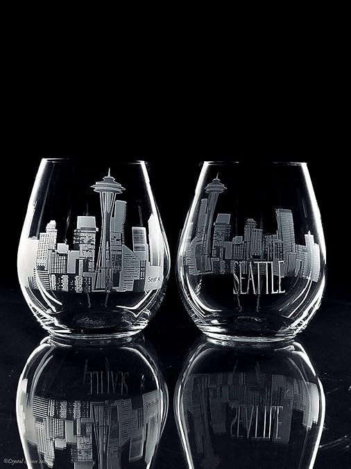 Two clear stemless wine glasses with etched Seattle skyline designs, shown on a reflective black surface against a black background.
