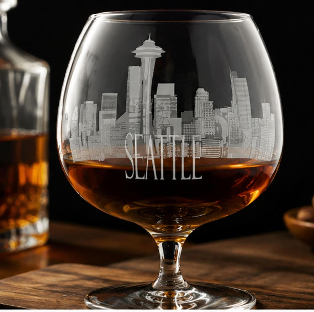 A clear glass snifter tumbler with an etched Seattle skyline and the word 'SEATTLE' on the front, filled with amber liquor, set on a wooden surface with a blurred bottle in the background.